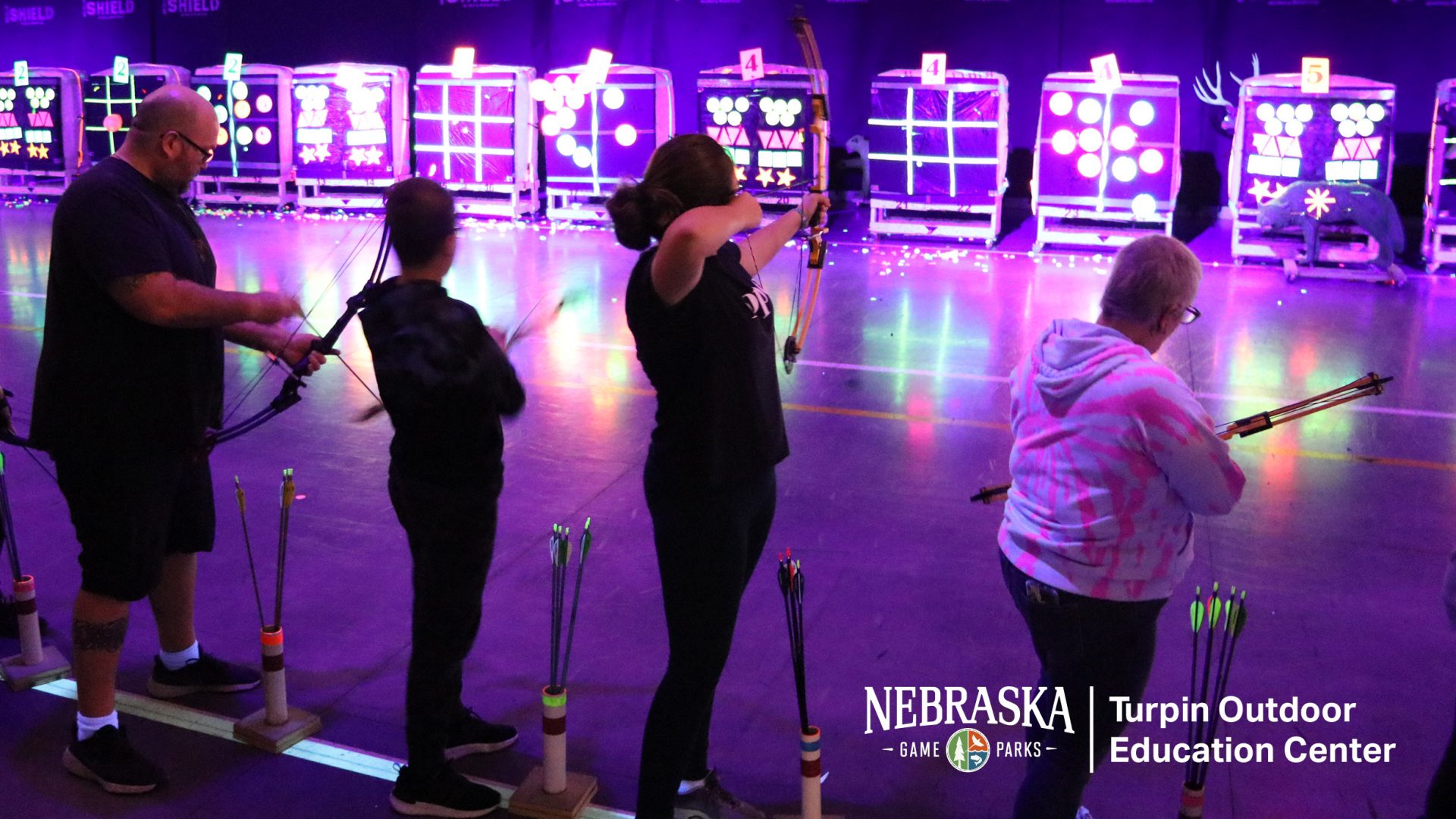 Family using bows on indoor archery range with purple lighting and glowing targets