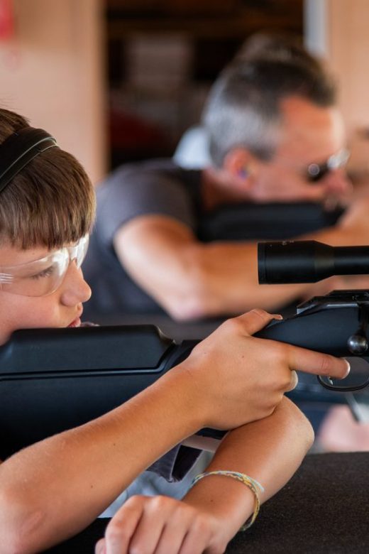 A boy wearing protective gear shoots a rifle at a shooting range.