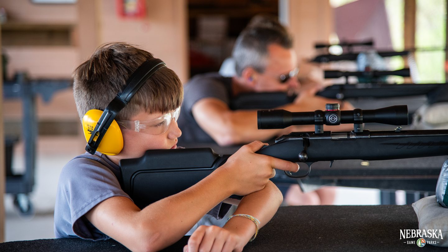A boy wearing protective gear shoots a rifle at a shooting range.