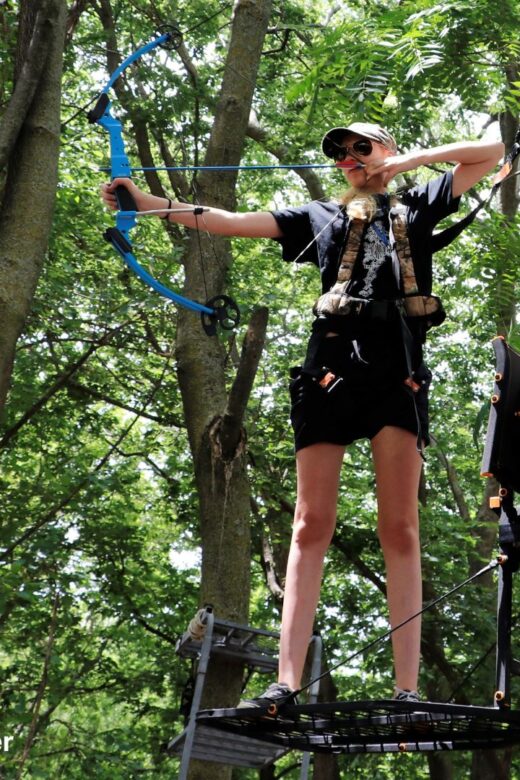 A man shoots archery outdoors from a tree stand.
