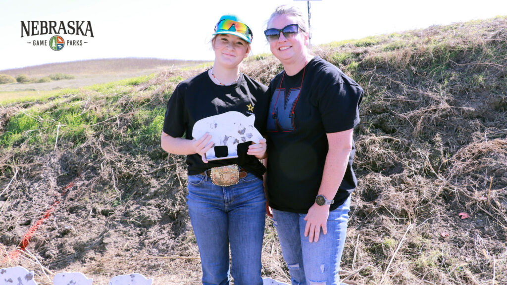 Mother and teenage daughter standing smiling together outdoors in front of grassy hill. Daughter holding white metal pig target