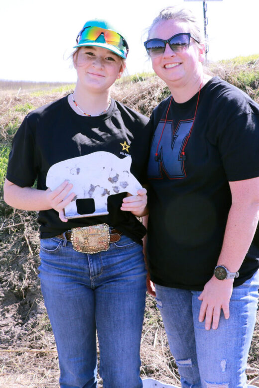 Mother and teenage daughter standing smiling together outdoors in front of grassy hill. Daughter holding white metal pig target